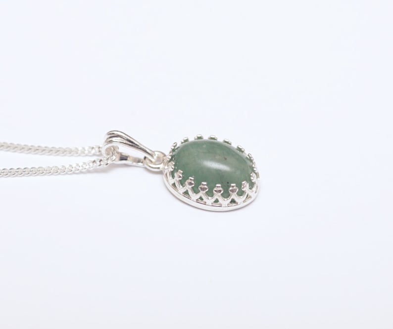 Puede incluir: Un collar con colgante de plata con una piedra preciosa ovalada de color verde claro. La piedra preciosa est&aacute; engastada en un bisel decorativo de plata. La cadena del collar es una delicada cadena de plata.