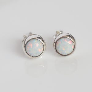 May include: Two stud earrings with round, iridescent opal stones set in silver metal. The opals show white, pink, and orange colours. The earrings have a simple, elegant design.
