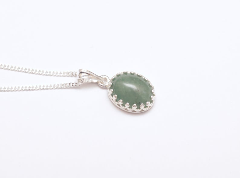 Puede incluir: Collar de plata con colgante. El colgante tiene una piedra preciosa ovalada de color verde claro engastada en un bisel decorativo de plata. La cadena del collar es una fina cadena de plata.