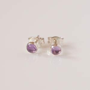 May include: A pair of stud earrings featuring round, polished purple amethyst gemstones set in silver metal settings. Each earring has a smooth, circular cabochon stone held by a simple metallic base, designed for pierced ears with a post back.