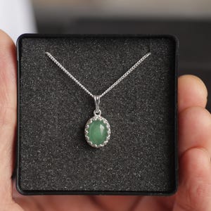 Puede incluir: Un collar de plata con un colgante ovalado de piedra verde. El colgante est&aacute; engastado en un marco de plata decorativo y cuelga de una delicada cadena. El collar se presenta en una caja negra con un interior texturizado.