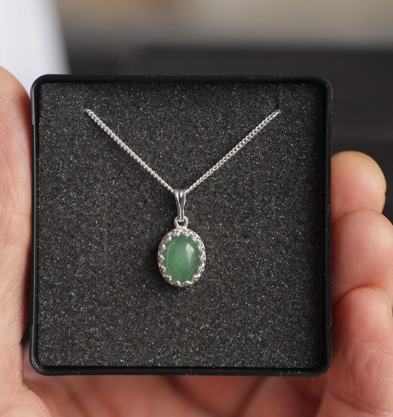 Puede incluir: Un collar de plata con un colgante ovalado de piedra verde. El colgante est&aacute; engastado en un marco de plata decorativo y cuelga de una delicada cadena. El collar se presenta en una caja negra con un interior texturizado.
