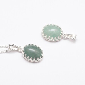 Puede incluir: Dos colgantes ovalados con una piedra verde claro engastada en un bisel plateado con forma de corona. Un colgante est&aacute; en una cadena plateada. Los colgantes se muestran sobre un fondo blanco, mostrando el dise&ntilde;o de la joyer&iacute;a.