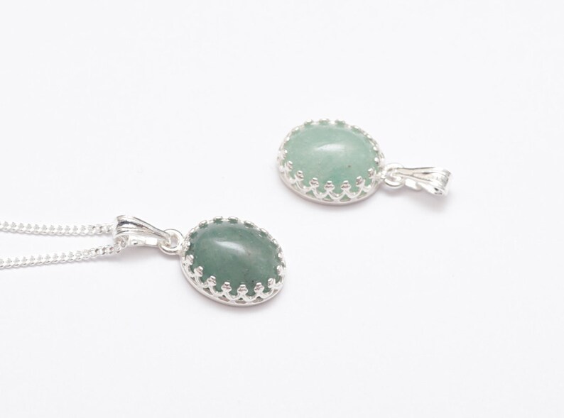 Puede incluir: Dos colgantes ovalados con una piedra verde claro engastada en un bisel plateado con forma de corona. Un colgante est&aacute; en una cadena plateada. Los colgantes se muestran sobre un fondo blanco, mostrando el dise&ntilde;o de la joyer&iacute;a.