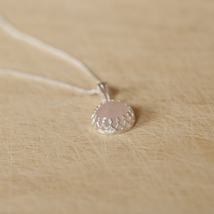 Op de afbeelding: Een zilveren hangerketting met een ronde, lichtroze edelsteen. De edelsteen is gezet in een decoratieve, kroonachtige zilveren zetting. De ketting is dun en zilverkleurig. De achtergrond is een lichtbruin, getextureerd oppervlak.
