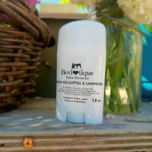 Op de afbeelding: Een witte deodorantstick met de tekst "Bovi tique" en "Lemon Eucalyptus & Lavender". Het etiket vermeldt ook ingrediënten en het gewicht van 51 gram. De achtergrond bevat bloemen en een rieten mand.