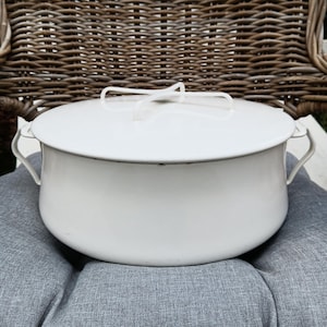 May include: A white enamelware pot with a lid, featuring two side handles and a top handle. The pot is set on a gray cushion, with a wicker chair in the background. The pot has a classic, vintage design.