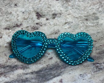 May include: Turquoise heart-shaped sunglasses with a sparkling rhinestone border. The lenses are a matching turquoise color. The sunglasses are a fun and fashionable accessory.