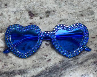 May include: Blue heart-shaped sunglasses with a sparkling rhinestone border. The lenses are a deep blue, and the frames are also blue. These fashion sunglasses are a fun accessory.