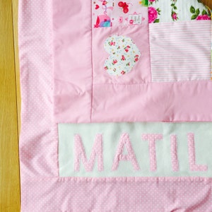 May include: A pink and white patchwork baby blanket with a heart-shaped applique and the name "MATIL" embroidered on the bottom.