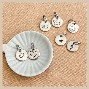 May include: A collection of seven silver charm pendants with various designs, including a heart, a star, a musical note, a cat, a dog, and two hearts. The charms are displayed on a white plate and a beige surface.