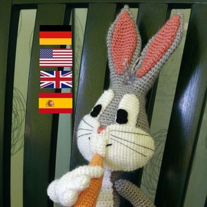 Könnte beinhalten: Ein gehäkeltes Plüschtier von Bugs Bunny, das eine Karotte hält. Das Spielzeug ist grau und weiß mit rosa Ohren und schwarzen Augen. Die Karotte ist orange mit grünen Blättern. Das Spielzeug sitzt auf einem schwarzen Stuhl mit einem grünen Kissen.