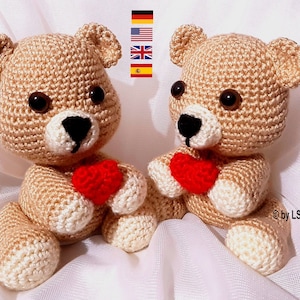 May include: Two crocheted teddy bears holding red hearts. The bears are beige with brown eyes and black noses. They are sitting on a white surface.