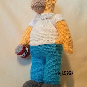 May include: A handmade crochet doll of Homer Simpson, featuring a yellow skin tone, white shirt, blue pants, and gray shoes. The doll holds a red and white object in its left hand. The doll is approximately 18 inches tall.