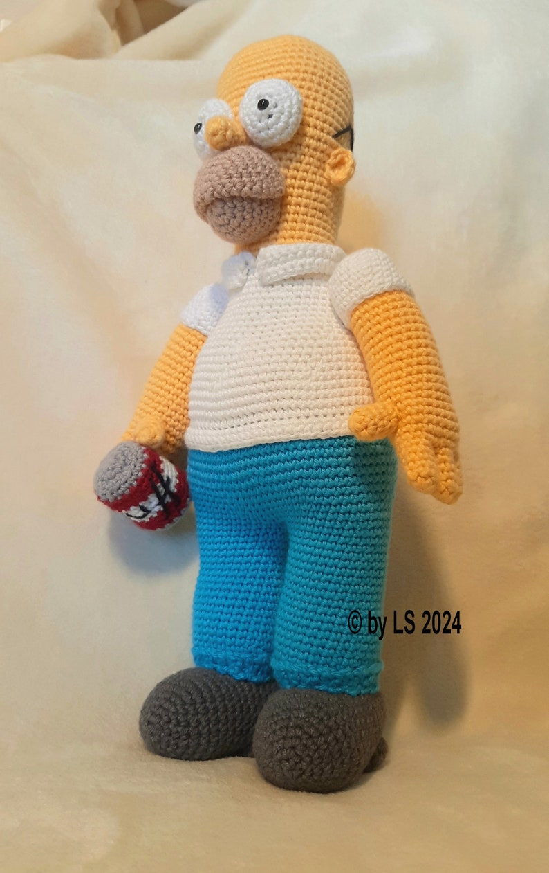 May include: A handmade crochet doll of Homer Simpson, featuring a yellow skin tone, white shirt, blue pants, and gray shoes. The doll holds a red and white object in its left hand. The doll is approximately 18 inches tall.