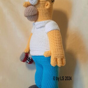May include: Crocheted Homer Simpson doll. The doll features a yellow head, arms, and hands, a white shirt, blue pants, and gray shoes. It holds a red, white, and gray object. The doll is handmade and has black glasses.