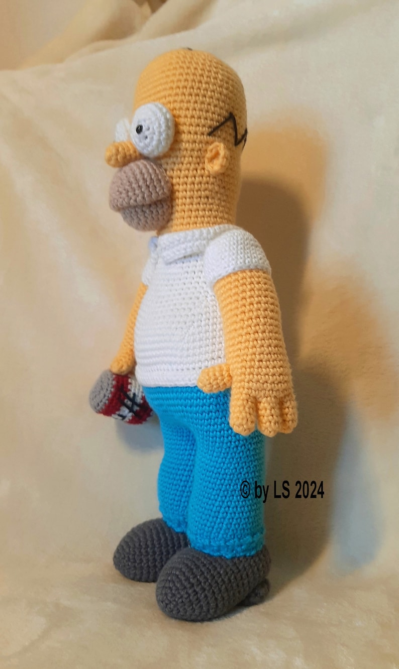 May include: Crocheted Homer Simpson doll. The doll features a yellow head, arms, and hands, a white shirt, blue pants, and gray shoes. It holds a red, white, and gray object. The doll is handmade and has black glasses.