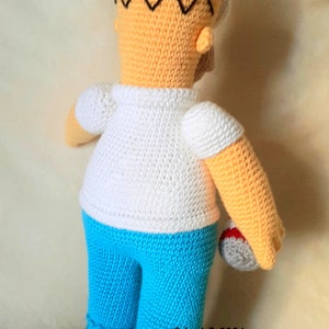 May include: Crocheted figure of a cartoon character, featuring a yellow head with black hair, a white shirt, and blue pants. The figure is holding a gray ball with red accents. The figure is handmade.