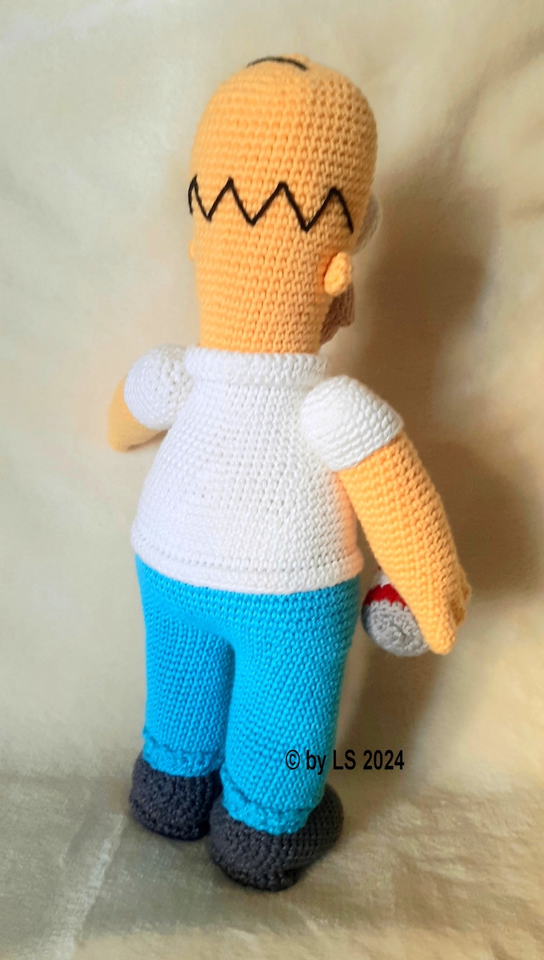 May include: Crocheted figure of a cartoon character, featuring a yellow head with black hair, a white shirt, and blue pants. The figure is holding a gray ball with red accents. The figure is handmade.