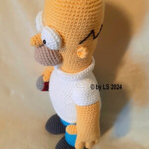 May include: Handmade crochet doll of Homer Simpson, featuring a yellow head, white shirt, and blue pants. The doll has black hair, glasses, and a detailed face. The doll is approximately 8 inches tall.