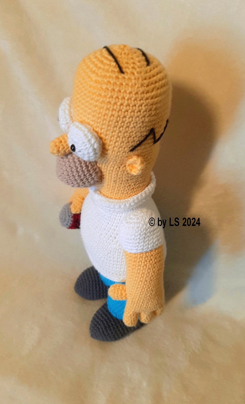 May include: Handmade crochet doll of Homer Simpson, featuring a yellow head, white shirt, and blue pants. The doll has black hair, glasses, and a detailed face. The doll is approximately 8 inches tall.