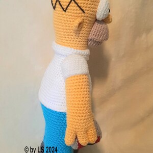 May include: A handmade crochet figure of Homer Simpson, featuring a yellow head with black hair, a white shirt, blue pants, and gray shoes. The figure is approximately 12 inches tall and is a side view.
