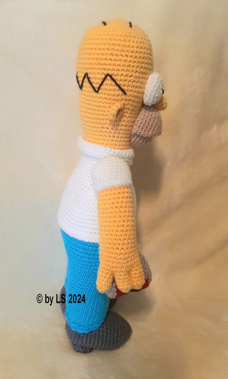 May include: A handmade crochet figure of Homer Simpson, featuring a yellow head with black hair, a white shirt, blue pants, and gray shoes. The figure is approximately 12 inches tall and is a side view.