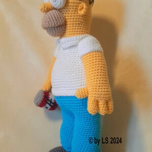 May include: Crocheted figure of Homer Simpson, featuring a yellow head, white shirt, blue pants, and gray shoes. The figure holds a red and white object. The copyright symbol and the text "by LS 2024" are visible on the right side.