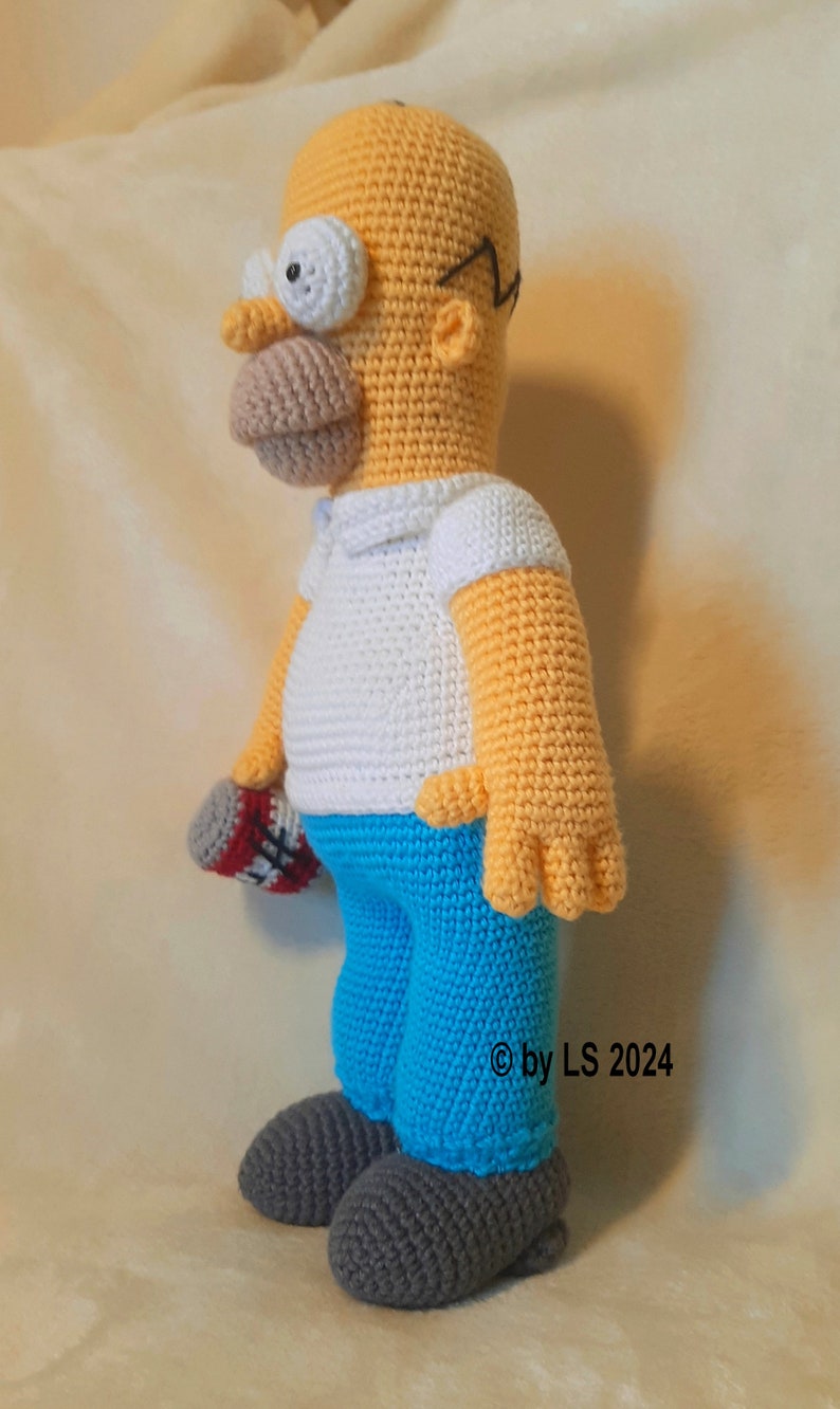May include: Crocheted figure of Homer Simpson, featuring a yellow head, white shirt, blue pants, and gray shoes. The figure holds a red and white object. The copyright symbol and the text "by LS 2024" are visible on the right side.