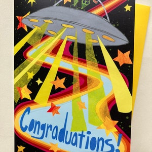 Op de afbeelding: Een felicitatiekaart met een kleurrijke illustratie van een UFO met een groene alien die een afstudeerpet draagt en een vredesteken maakt. De UFO vliegt door een regenboogkleurige lichtspoor met sterren op de achtergrond. De tekst "Gefeliciteerd!" staat in blauw op een lichtblauwe achtergrond.