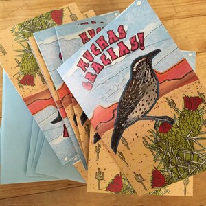 May include: A stack of greeting cards with light blue envelopes. The cards feature a desert scene with a bird, cactus, and the words "MUCHAS GRACIAS!" in pink. The background is a light tan color with cactus and fork illustrations.