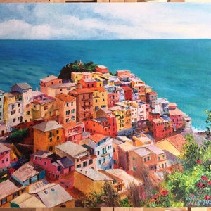 May include: A colourful painting of a seaside town with buildings clustered together on a cliff overlooking a turquoise ocean. The sky is blue with white clouds.