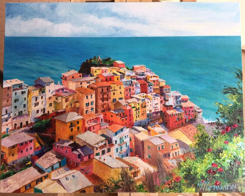 May include: A colourful painting of a seaside town with buildings clustered together on a cliff overlooking a turquoise ocean. The sky is blue with white clouds.