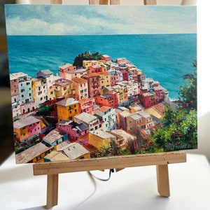 May include: A painting depicting a colourful Italian village perched on a hillside overlooking the sea. The village is painted in shades of yellow, orange, red, pink, and blue. The sea is a deep blue, and the sky is a light blue with white clouds. The painting is signed "M. Levant 21".