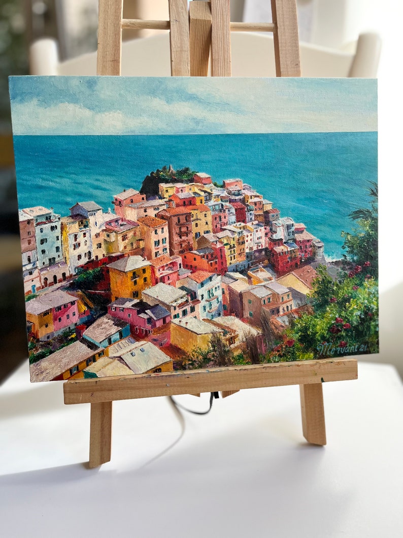 May include: A painting depicting a colourful Italian village perched on a hillside overlooking the sea. The village is painted in shades of yellow, orange, red, pink, and blue. The sea is a deep blue, and the sky is a light blue with white clouds. The painting is signed "M. Levant 21".