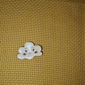 May include: A white cloud-shaped enamel pin with a smiling face and red cheeks. The pin has a silver-coloured outline. The pin is on a yellow cross-stitch fabric background.