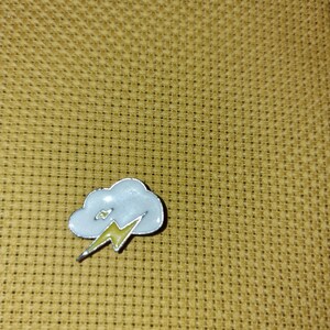 May include: A small enamel pin depicting a cloud and lightning bolt. The cloud is white with a silver border, and the lightning is yellow. The pin is set against a textured, golden-yellow background.