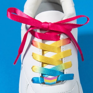Könnte beinhalten: Weißer Sneaker mit Regenbogen-Schnürsenkeln und einem passenden Schnürsenkelverschluss. Die Schnürsenkel wechseln von Pink zu Gelb zu Blau. Der Text "MATCHING LACE LOCK + SHOELACE SET" steht unten.
