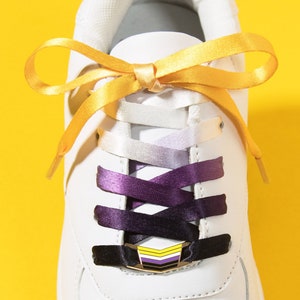 May include: White sneaker with a gold bow and non-binary pride shoelaces. The laces transition from white to purple and black, with a matching lace lock. The text "MATCHING LACE LOCK + SHOELACE SET" is at the bottom.