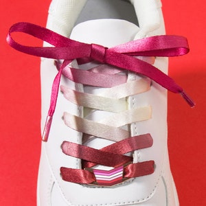 May include: White sneaker with pink and white gradient shoelaces tied in a bow. The shoelaces are a mix of pink shades. The text at the bottom reads "Matching Lace Lock + Shoelace Set" on a purple background.