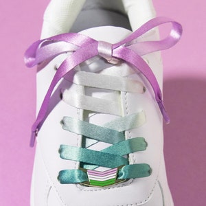 May include: White sneaker with gradient teal and purple shoelaces tied in a bow. The laces transition from white to teal and purple. The text "MATCHING LACE LOCK + SHOELACE SET" is at the bottom of the image.