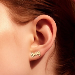 Op de afbeelding: Een close-up van iemands oor met een gouden oorbel waarop "they" in cursief staat.