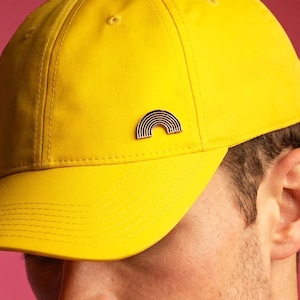 May include: Bright yellow baseball cap with a curved brim, featuring a small black and white rainbow emblem on the side. The cap is set against a pink background, and is worn with a pink t-shirt.