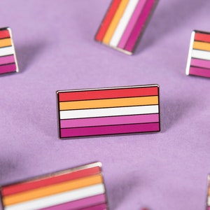 Op de afbeelding: Meerdere lesbische pride-vlagpinnen verspreid over een paars oppervlak. De pinnen zijn gemaakt van metaal en hebben de kleuren van de lesbische pride-vlag: oranje, roze, wit en lichtroze.