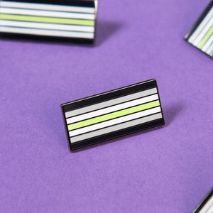 May include: Multiple black and white enamel pins with a thin green stripe. The pins are arranged on a purple background.