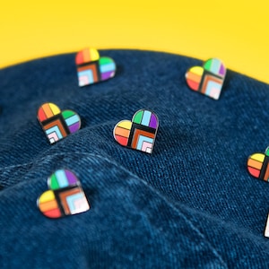 Puede incluir: Primer plano de una tela vaquera con alfileres en forma de corazón arcoíris. Los alfileres están esparcidos por la tela y todos miran en la misma dirección.