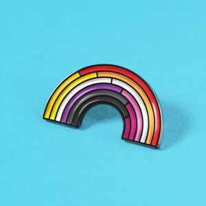 Op de afbeelding: Een regenboogvormige emaille pin met een zwarte omtrek en een verscheidenheid aan kleuren, waaronder geel, oranje, rood, paars en roze. De pin is geplaatst tegen een helderblauwe achtergrond.