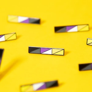 May include: Multiple yellow, purple, and black enamel pins with a geometric design. The pins are scattered on a yellow surface.