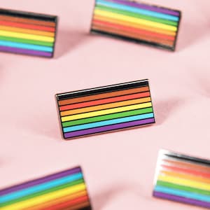 May include: A collection of rainbow pride flag pins on a pink background. The pins are all different sizes and shapes, but they all feature the same rainbow design.