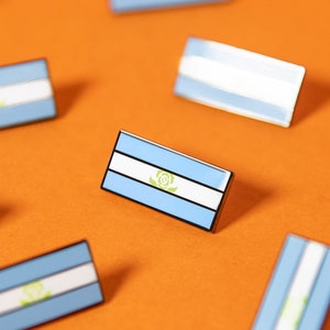May include: A collection of ten small blue and white enamel pins featuring the flag of Paraguay. The pins are scattered on a bright orange surface.